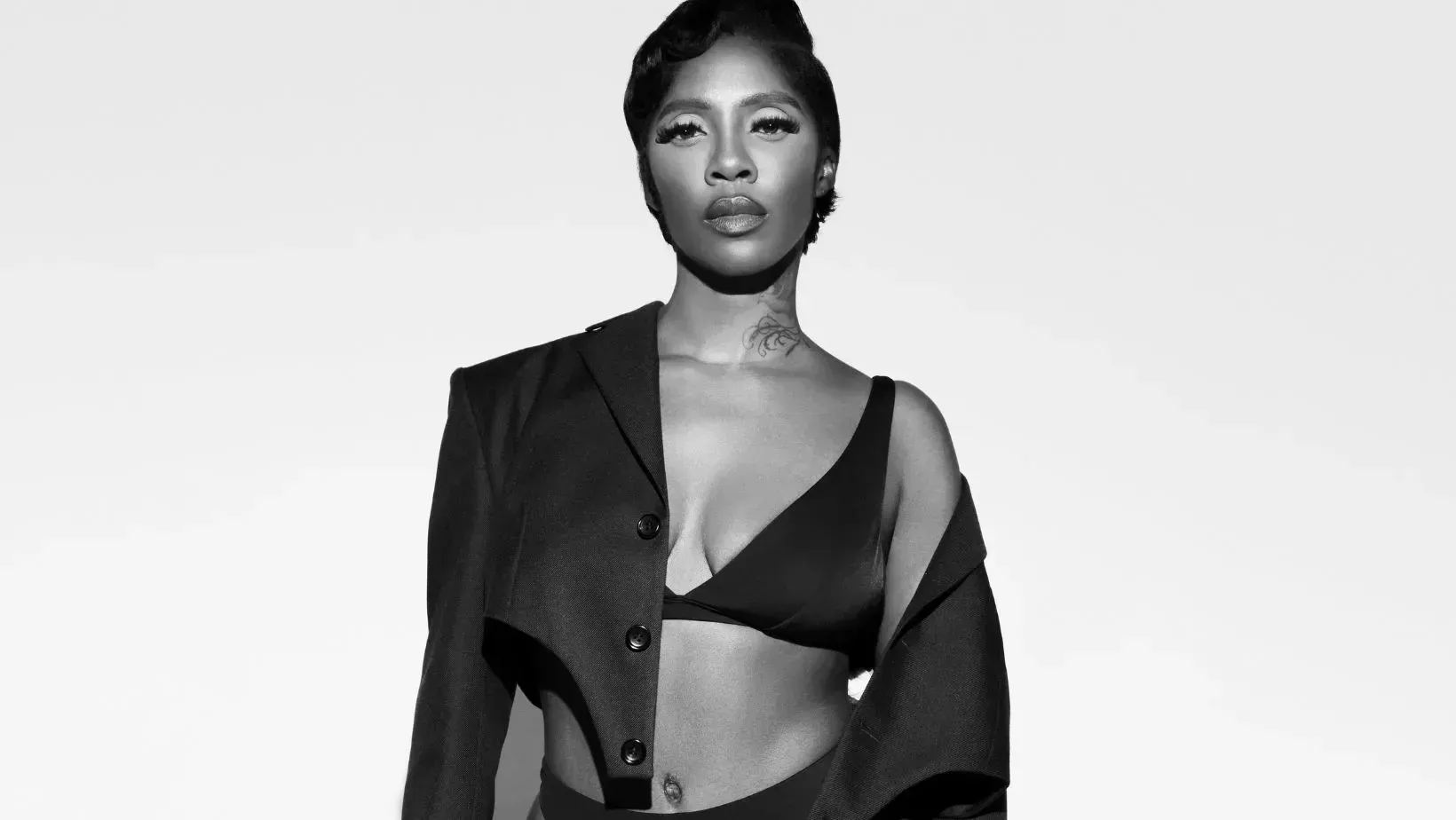 Black-and-white portrait of a model wearing an open jacket and bra, standing against a plain background