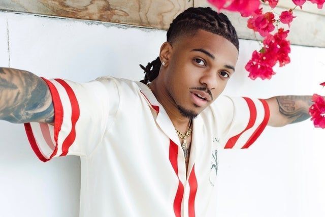 Man posing with arms outstretched in a white shirt with red stripes and pink flowers in the background