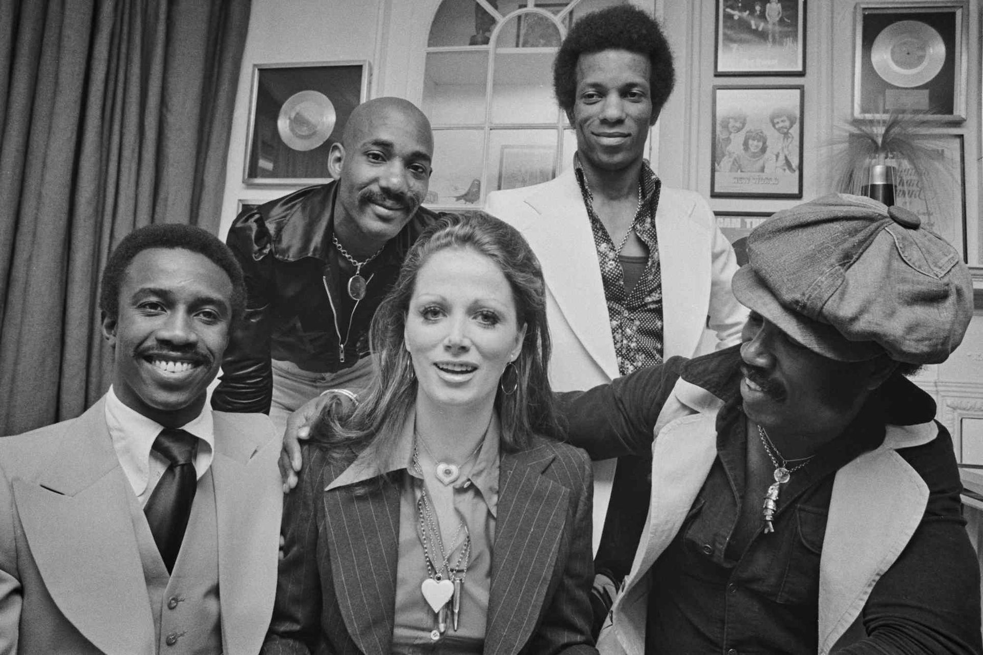 Five people pose indoors before curtains and framed records, smiling at the camera.
