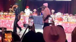 Performers on a brightly lit stage with pink floral décor, seen from an audience holding up phones.