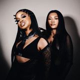 Two women posing in black outfits against a gray studio background, one wearing sunglasses and smiling.