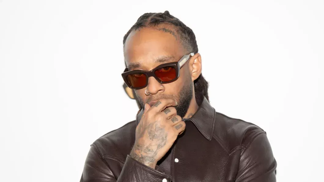 Man in sunglasses and a brown leather jacket poses against a white background, hand on chin.