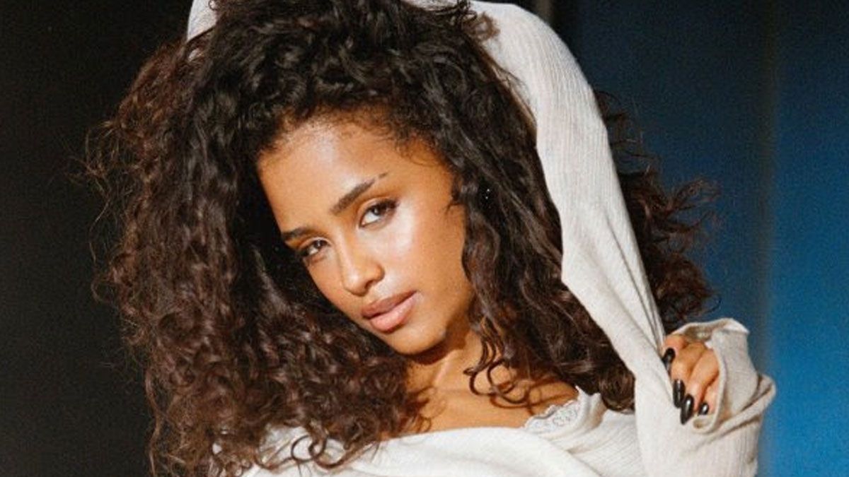 Portrait of a woman with curly hair in a white top, leaning against a gray backdrop, looking at the camera