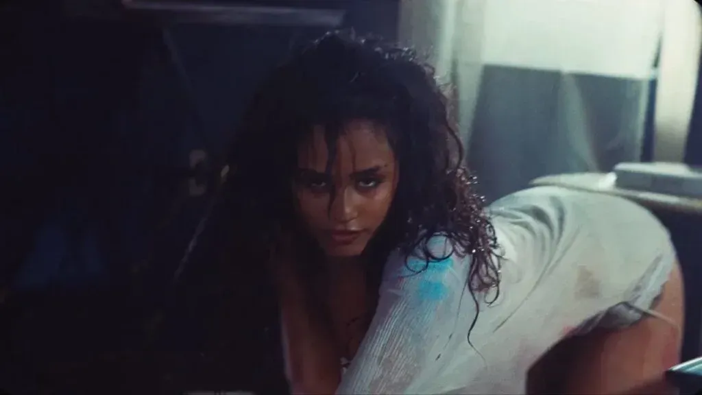 Person crouching in a dim room, staring intensely, with tousled dark hair and a light shirt.