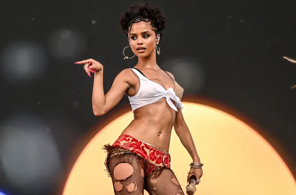 Singer performing onstage in a white top and red shorts, posing with a microphone against a glowing backdrop