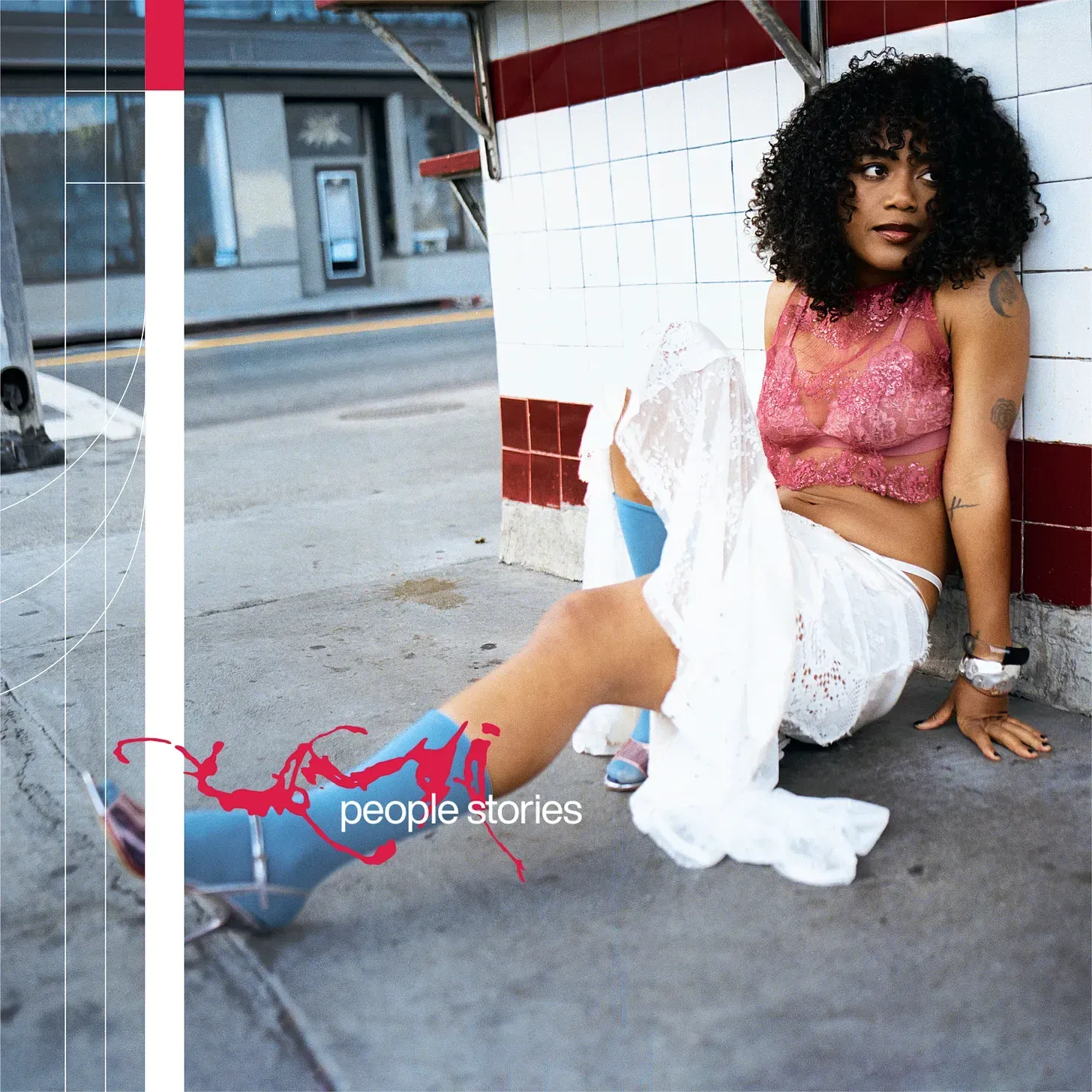 Woman in pink top and white pants sitting against a tiled wall, posing in an urban underpass.