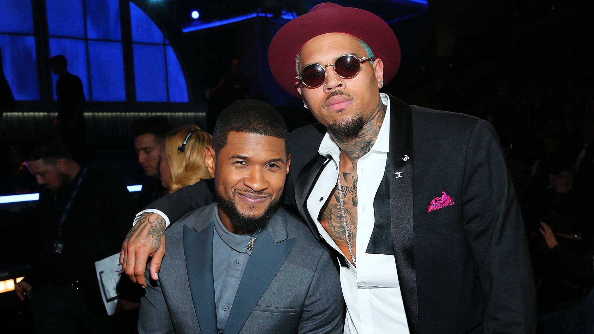 Two men posing together at a dim event, one in a red hat and sunglasses, the other in a dark blazer.