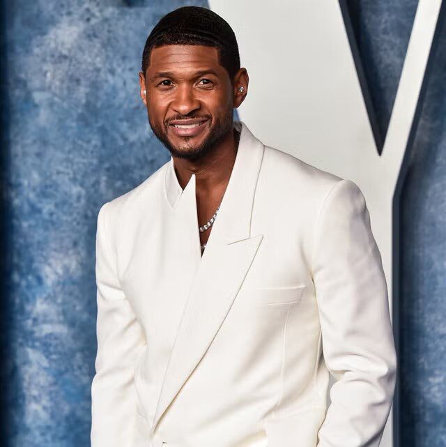 Man in a white suit smiling on a blue backdrop with a large letter V
