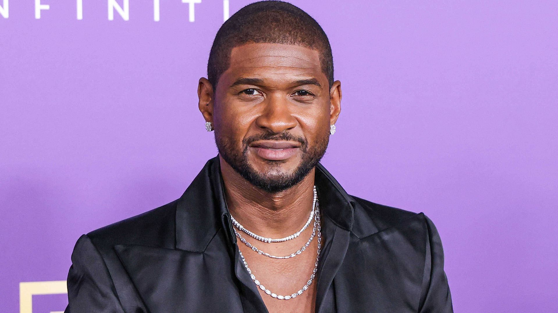 Man in a black jacket and layered necklaces posing before a purple event backdrop