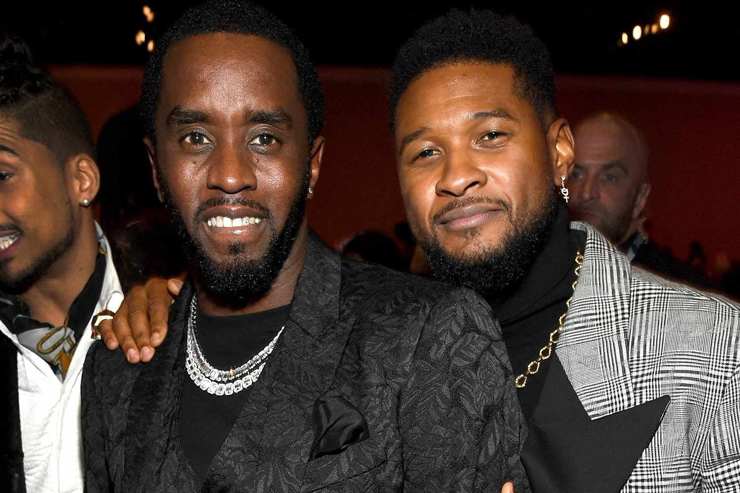 Two men smiling at a dimly lit event, wearing dark suits and chain necklaces