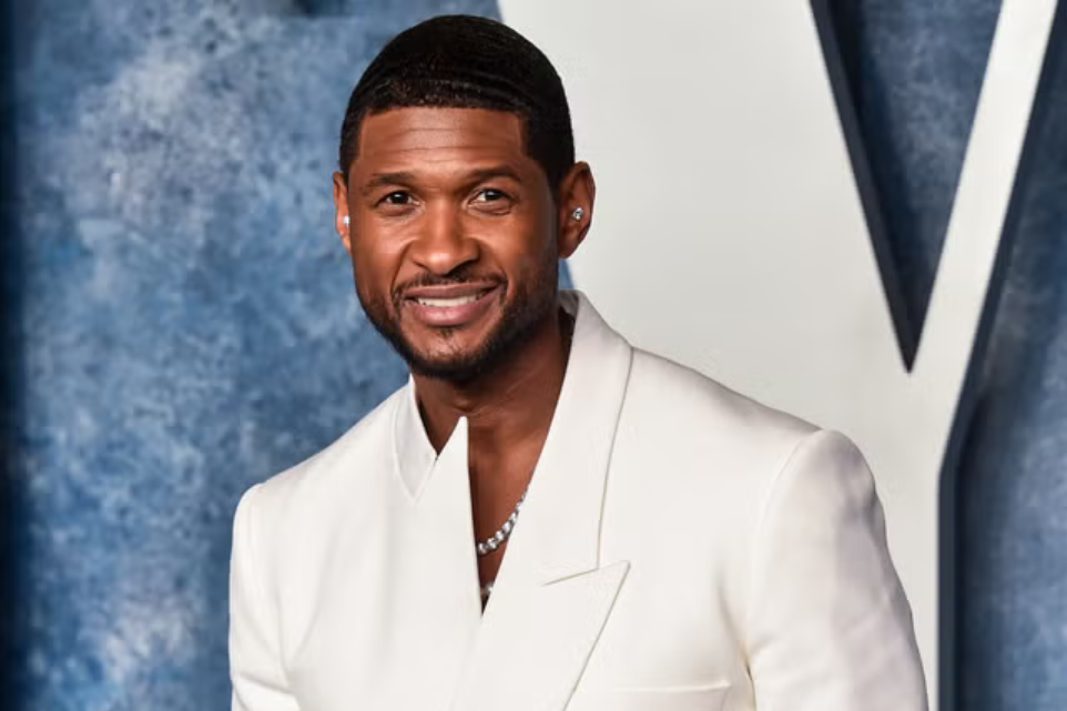Man in a white suit smiling on a red carpet with a large silver backdrop