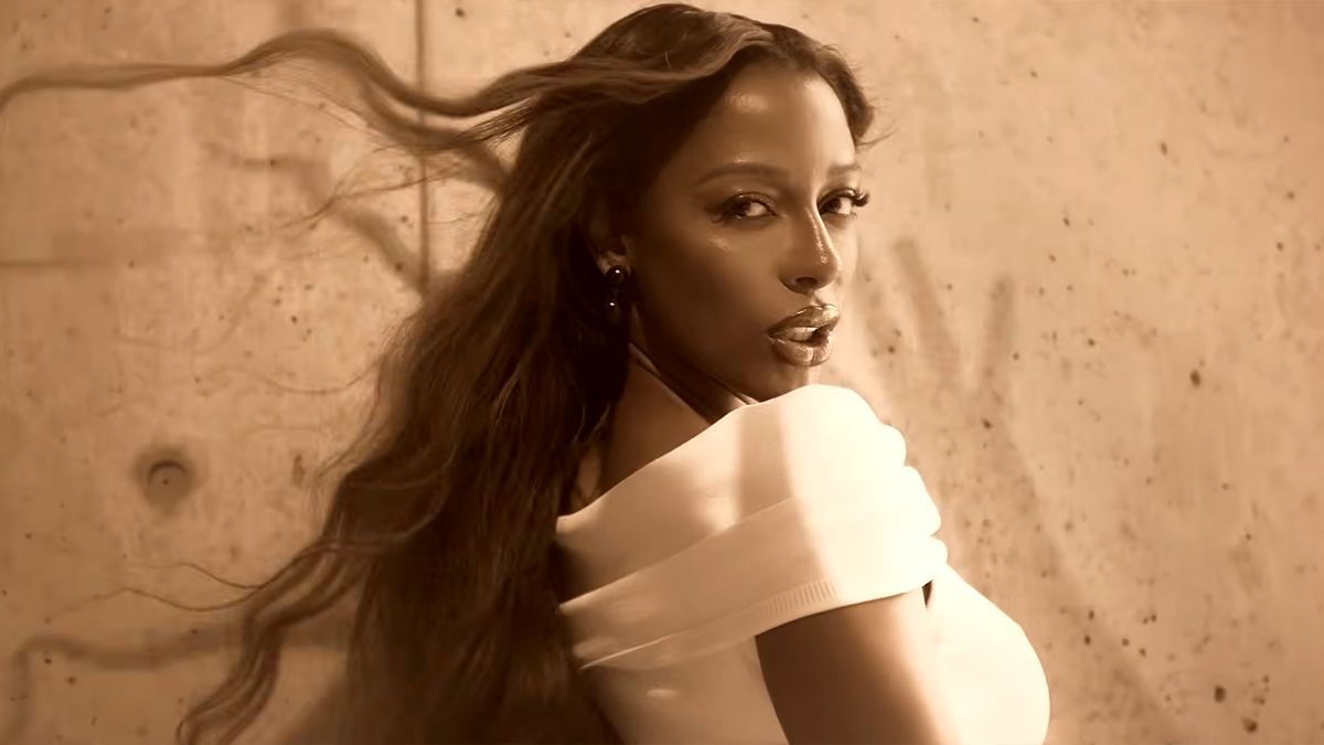 Woman with long hair turning toward the camera, wearing a white top against a warm-toned wall.