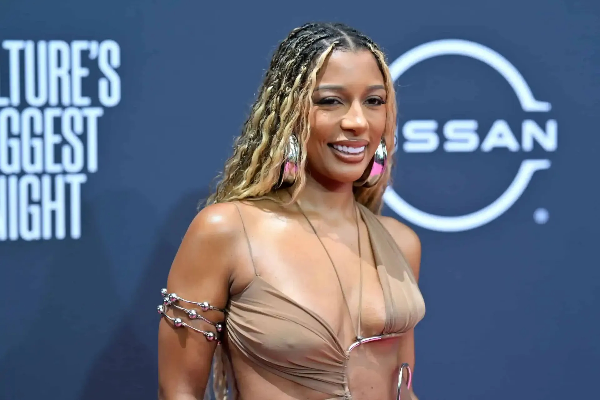 Woman in a sheer tan gown posing on a blue red-carpet backdrop with sponsor logos