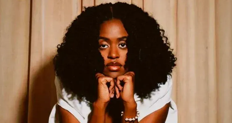 Portrait of a person with curly hair resting their chin on their hands against beige curtains