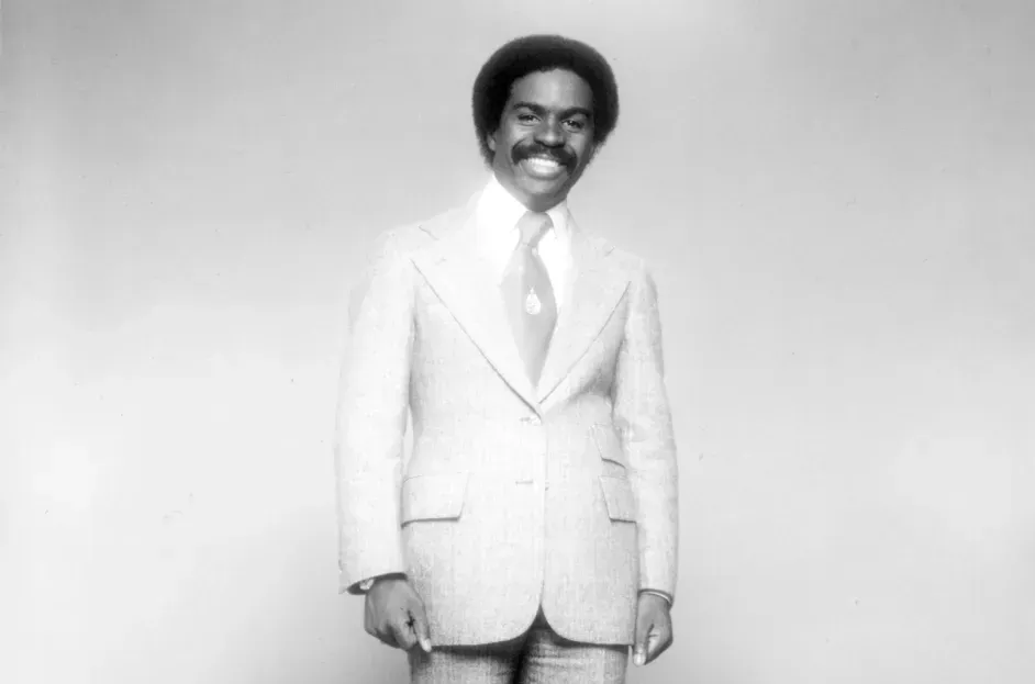 Man in a light suit and tie standing against a plain backdrop, smiling warmly