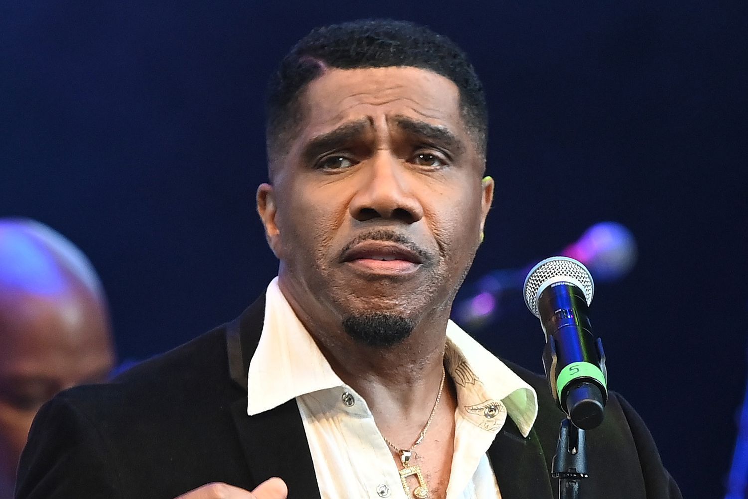 Man on stage speaking into a microphone, wearing a black jacket and white shirt under purple-blue lights