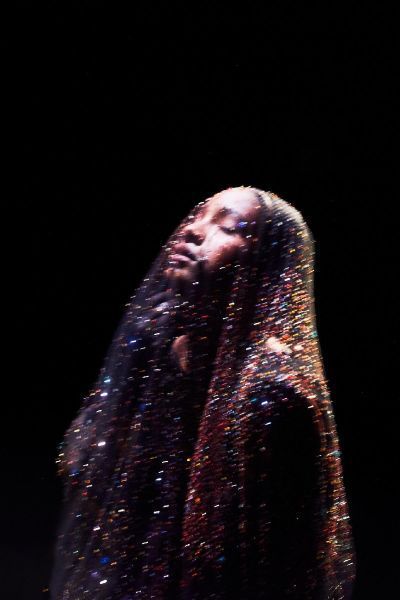 Person draped in glittering fabric against a black background, head tilted upward.
