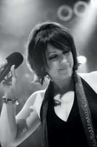 Woman singing into a microphone on stage, wearing a dark top and patterned guitar strap.