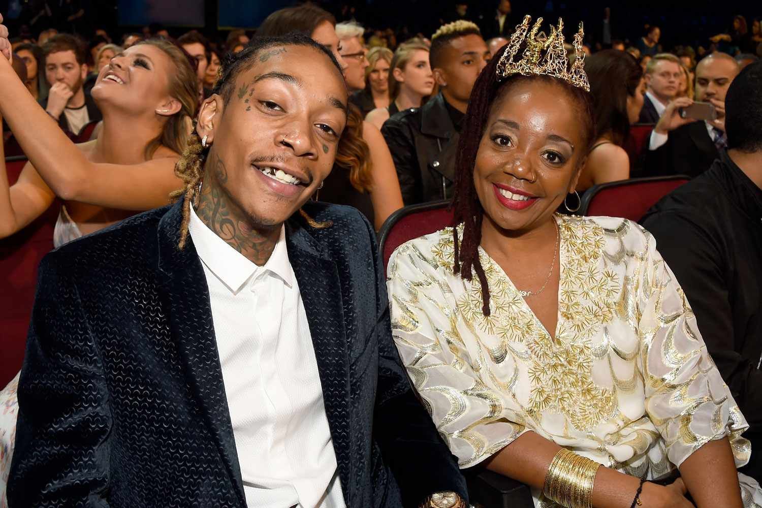 Two smiling people seated at a formal event, one in a dark suit and the other in a cream dress and crown