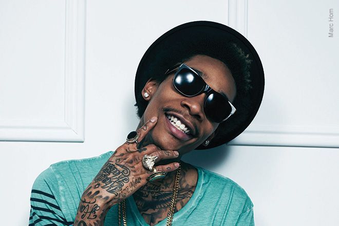 Smiling person in a black hat and sunglasses, with tattooed hands, against a light wall.