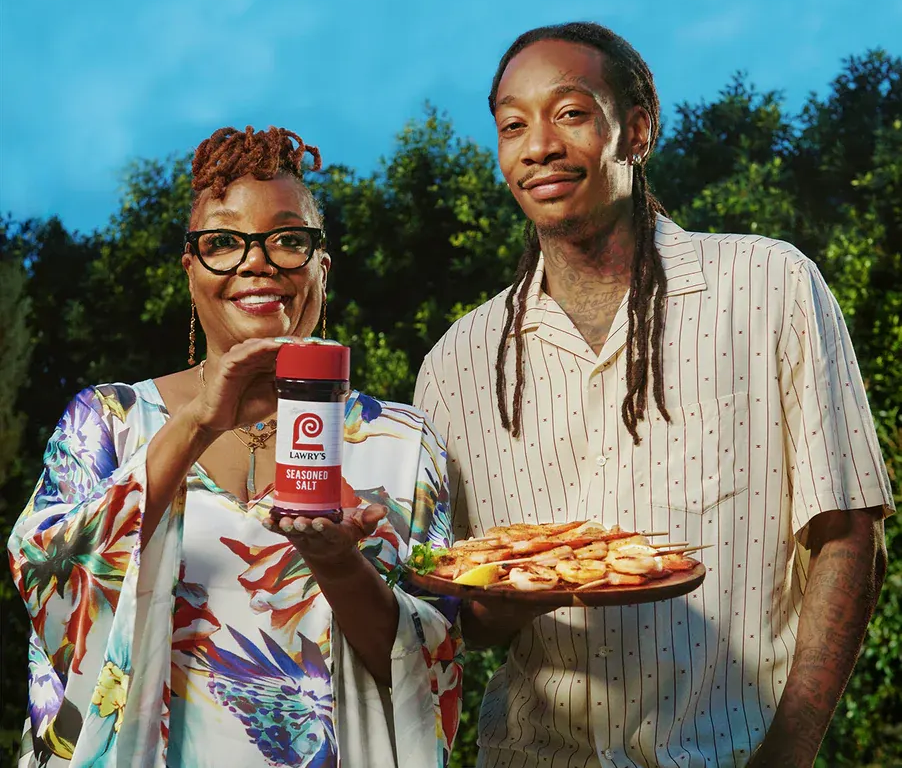 Two people outdoors holding a jar and a tray of food against a blue sky.