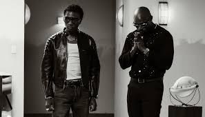 Two men standing indoors, one in a leather jacket and one in sunglasses with hands near his face.