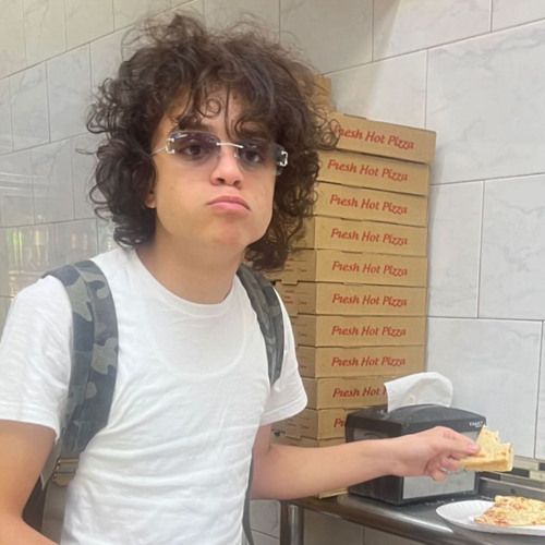 Person in a white T-shirt holding a slice of pizza in front of stacked pizza boxes, wearing sunglasses and a backpack.