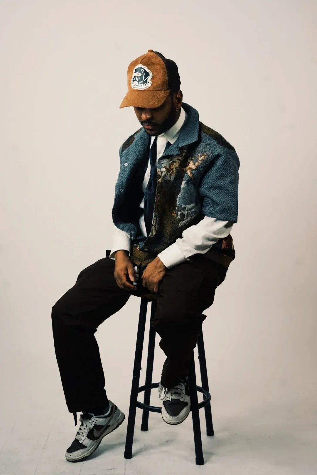 Person seated on a stool, wearing a tan cap, denim jacket, black pants, and gray sneakers against a plain backdrop.