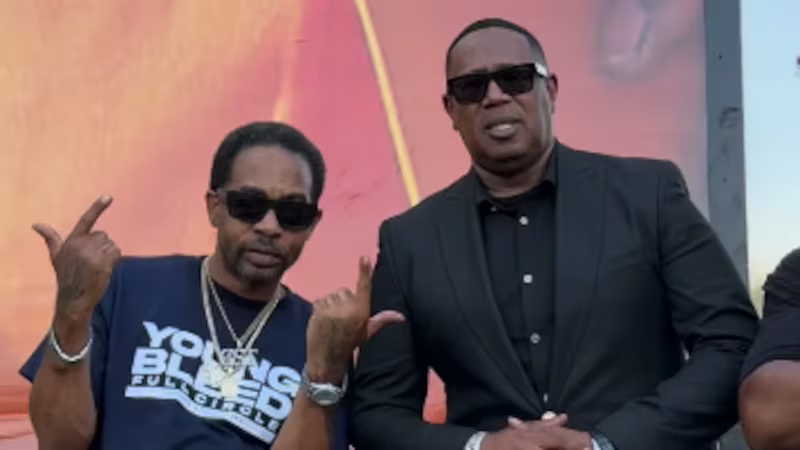 Two men in sunglasses pose outdoors, one making hand gestures, in front of a colorful backdrop.