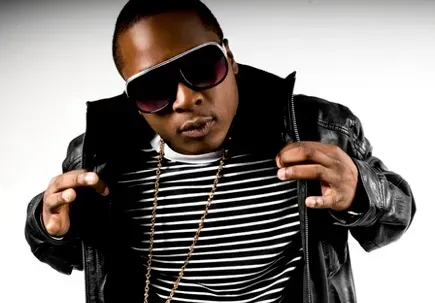 Smiling man in a white T-shirt, cap, sunglasses, and gold chain standing against a stone wall