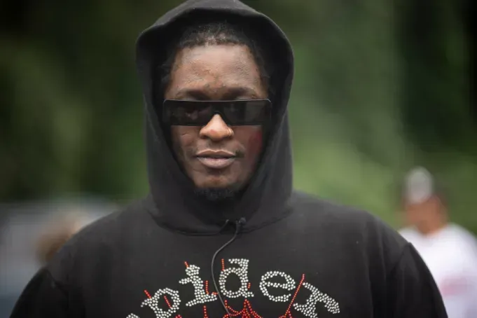 Person in a black hoodie and sunglasses, facing forward against a blurred outdoor background.