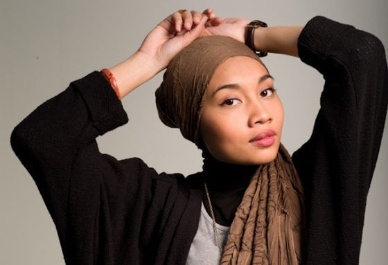 Woman in a brown headscarf posing with arms raised, wearing a black sweater on a neutral background