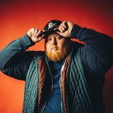 Red-bearded man in a teal jacket adjusting a black cap against a dark red background