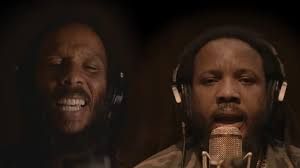 Two singers in a dark studio, one smiling and one at a microphone with headphones