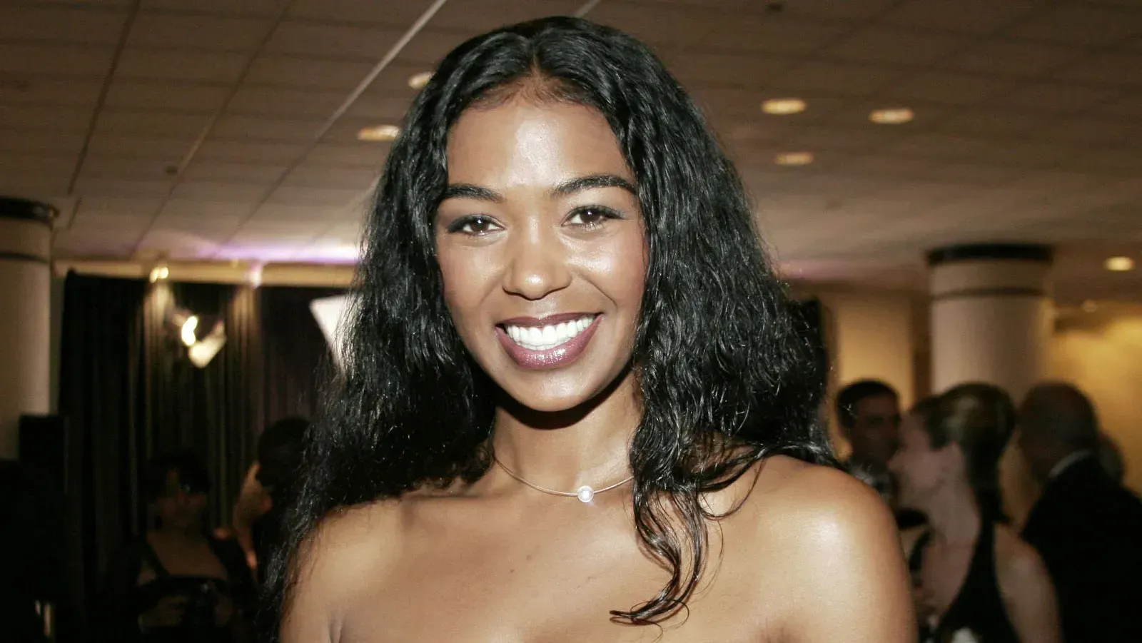 Smiling woman in a strapless black dress at an indoor event, with a softly lit crowd in the background