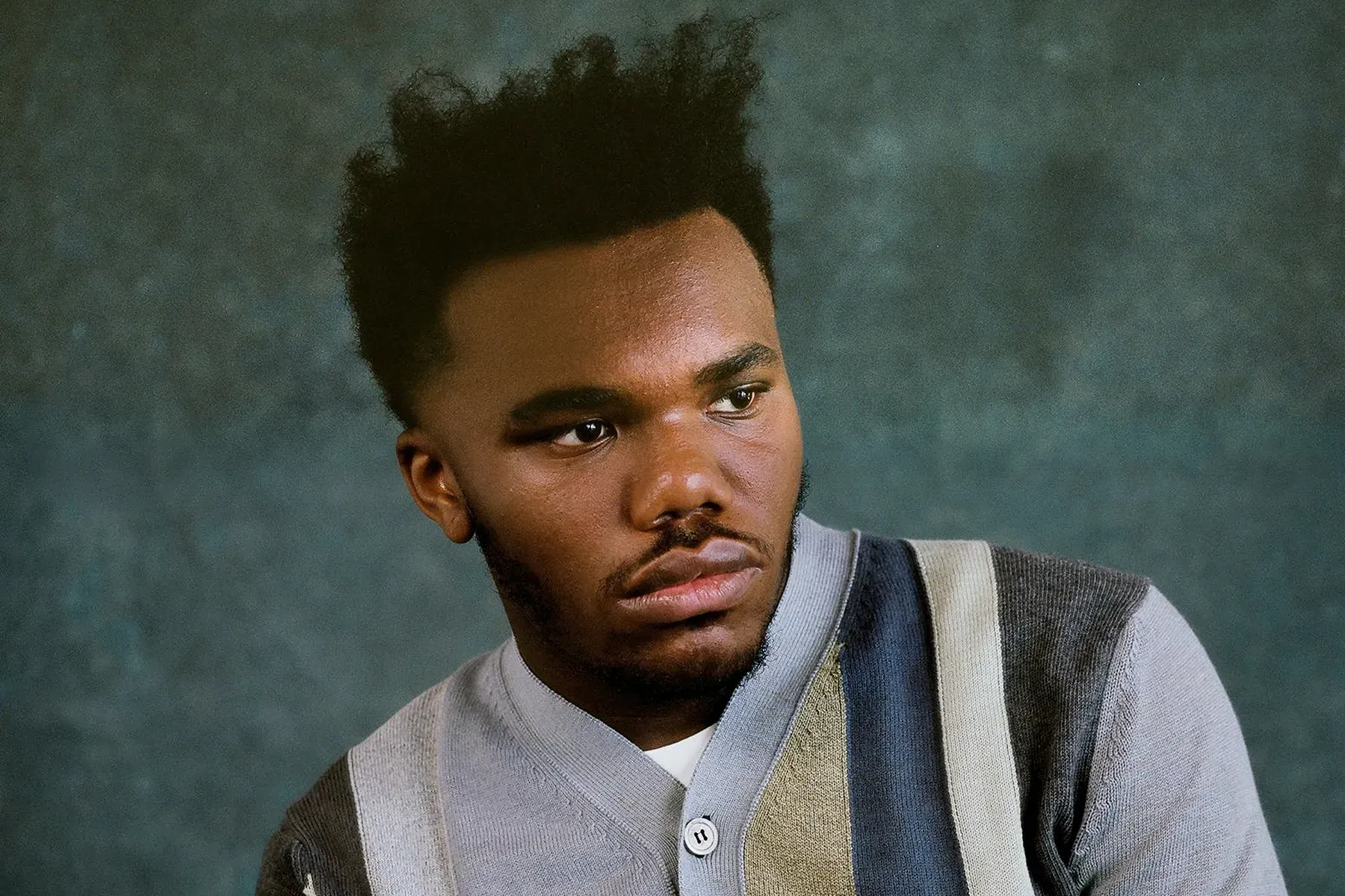 Portrait of a man in a gray shirt and striped sweater, looking to the side against a dark backdrop