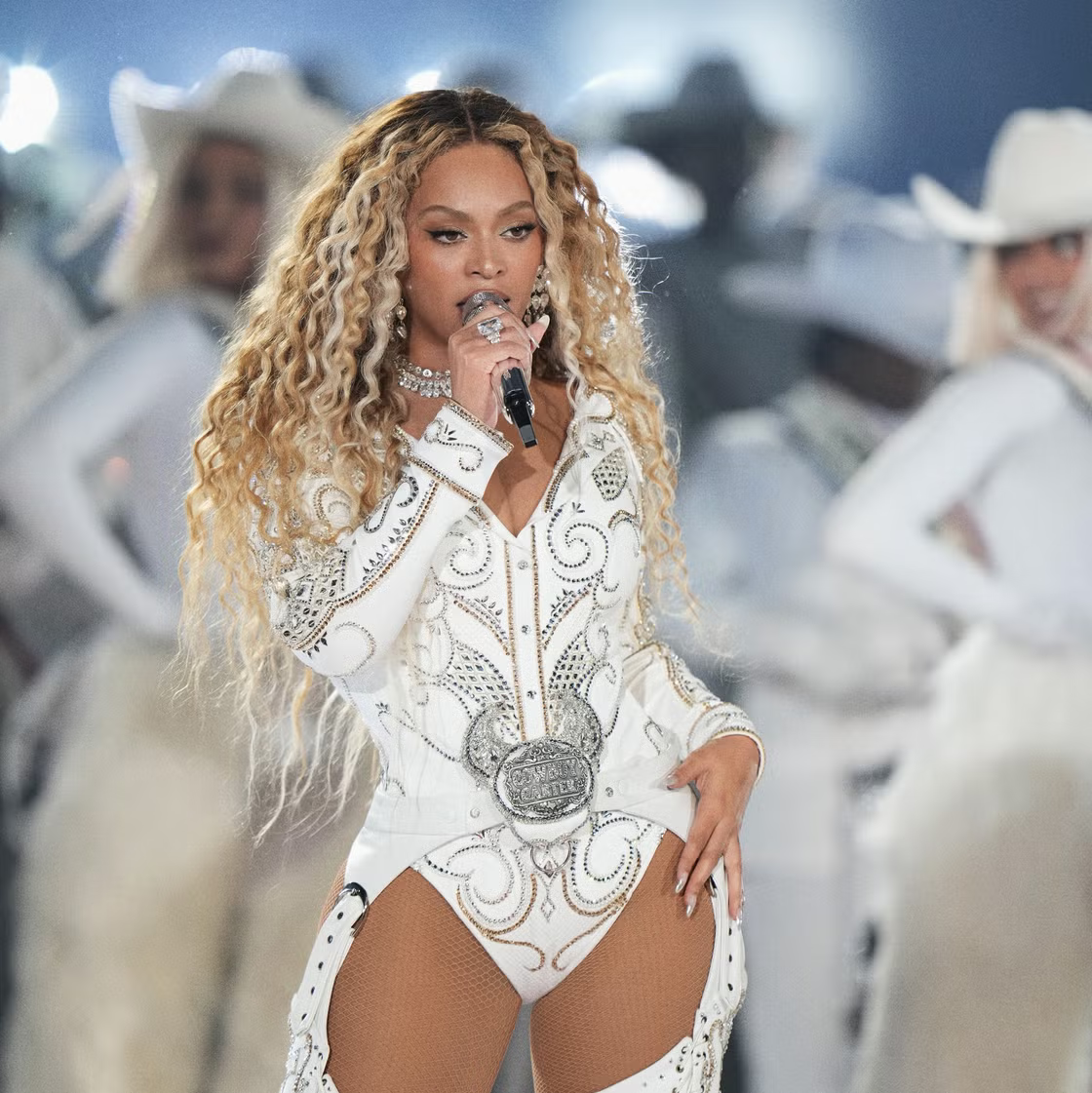 Singer performing onstage in a white jeweled bodysuit, holding a microphone with dancers behind her