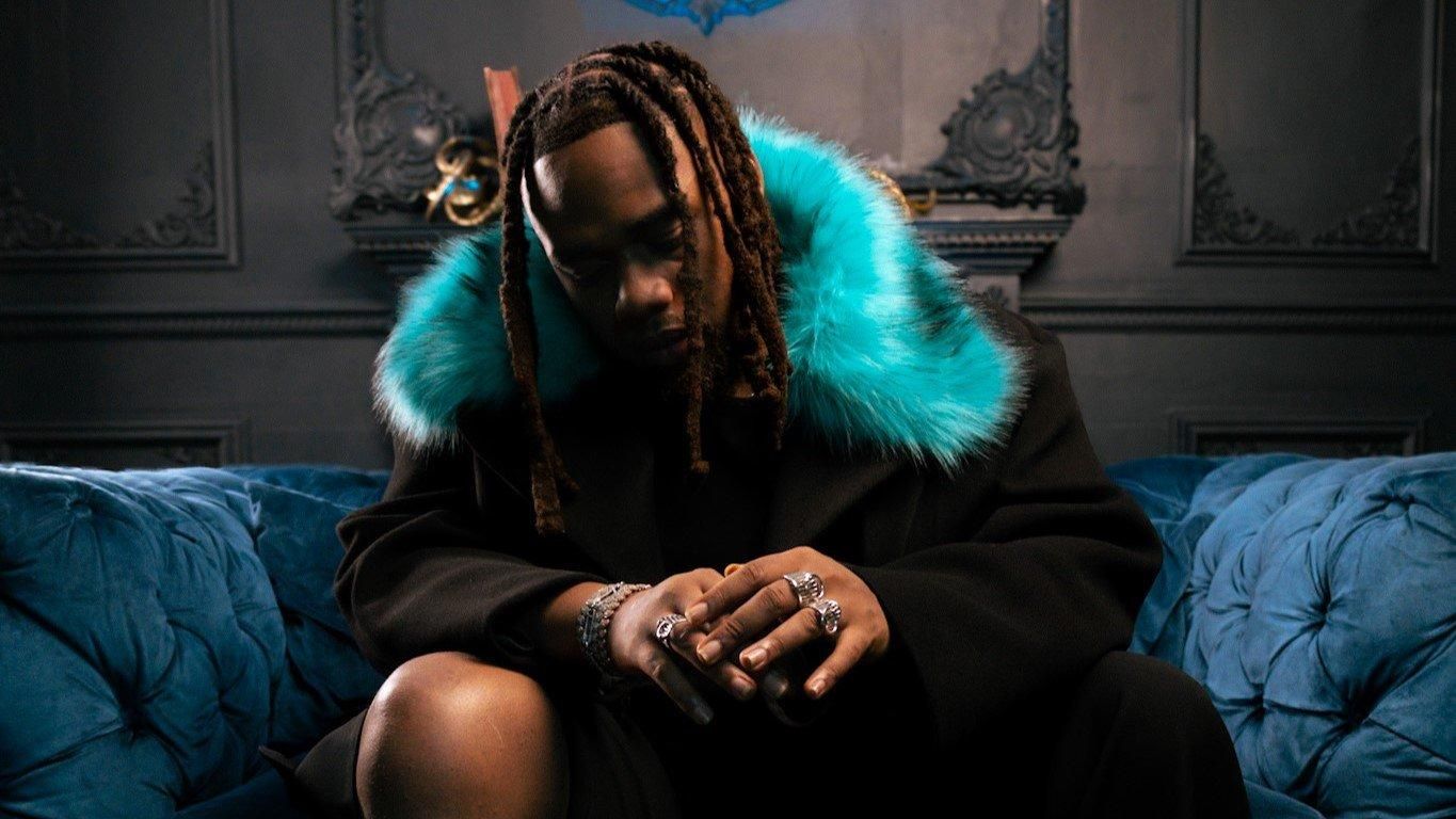 Person sitting on a blue couch in a dark room, wearing a black coat with a bright turquoise fur collar.