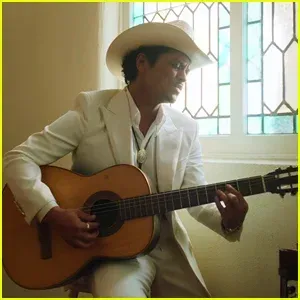 Man in a white suit and cowboy hat playing acoustic guitar by a sunlit window