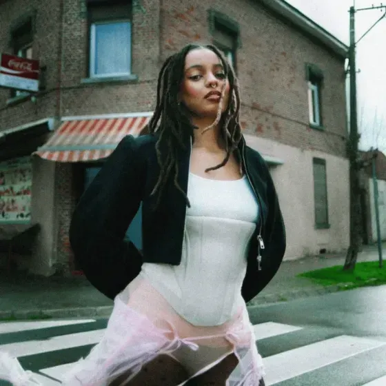 Person standing in a city street, wearing a black jacket and pink tulle skirt near a brick building.