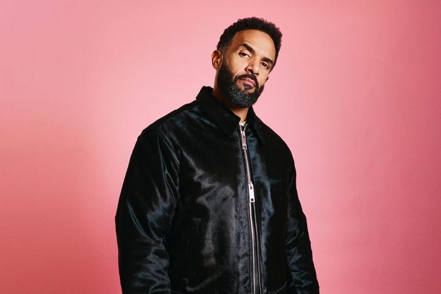 Man in a black jacket posing against a pink background, looking at the camera.