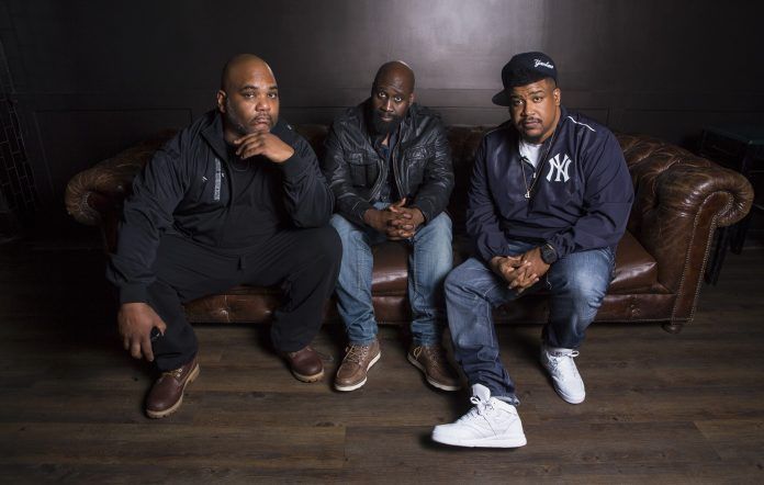 Three men seated on a brown leather couch in a dim room, posing for a photo.