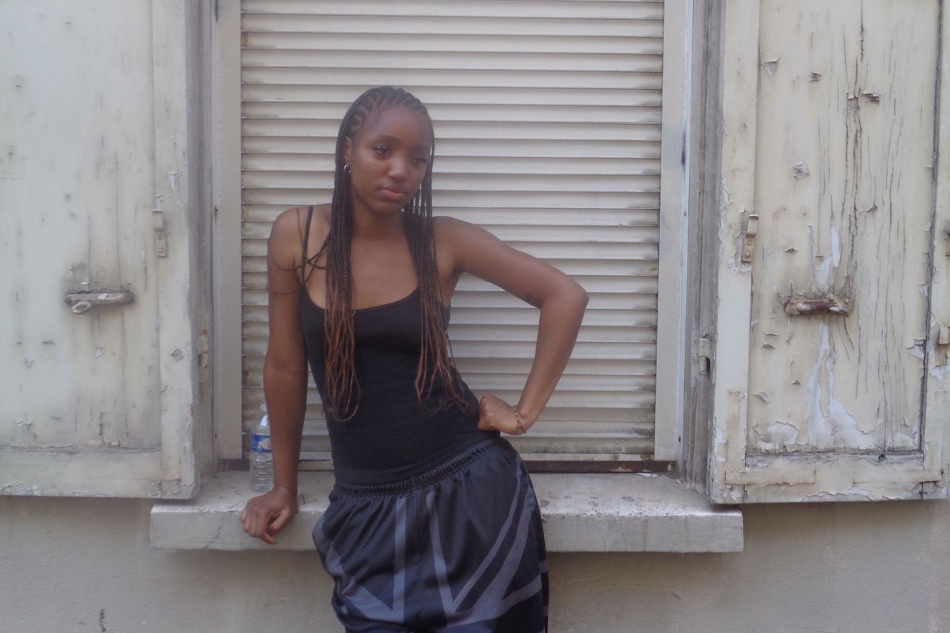 Person posing by a shuttered window, wearing a black top and shiny skirt.