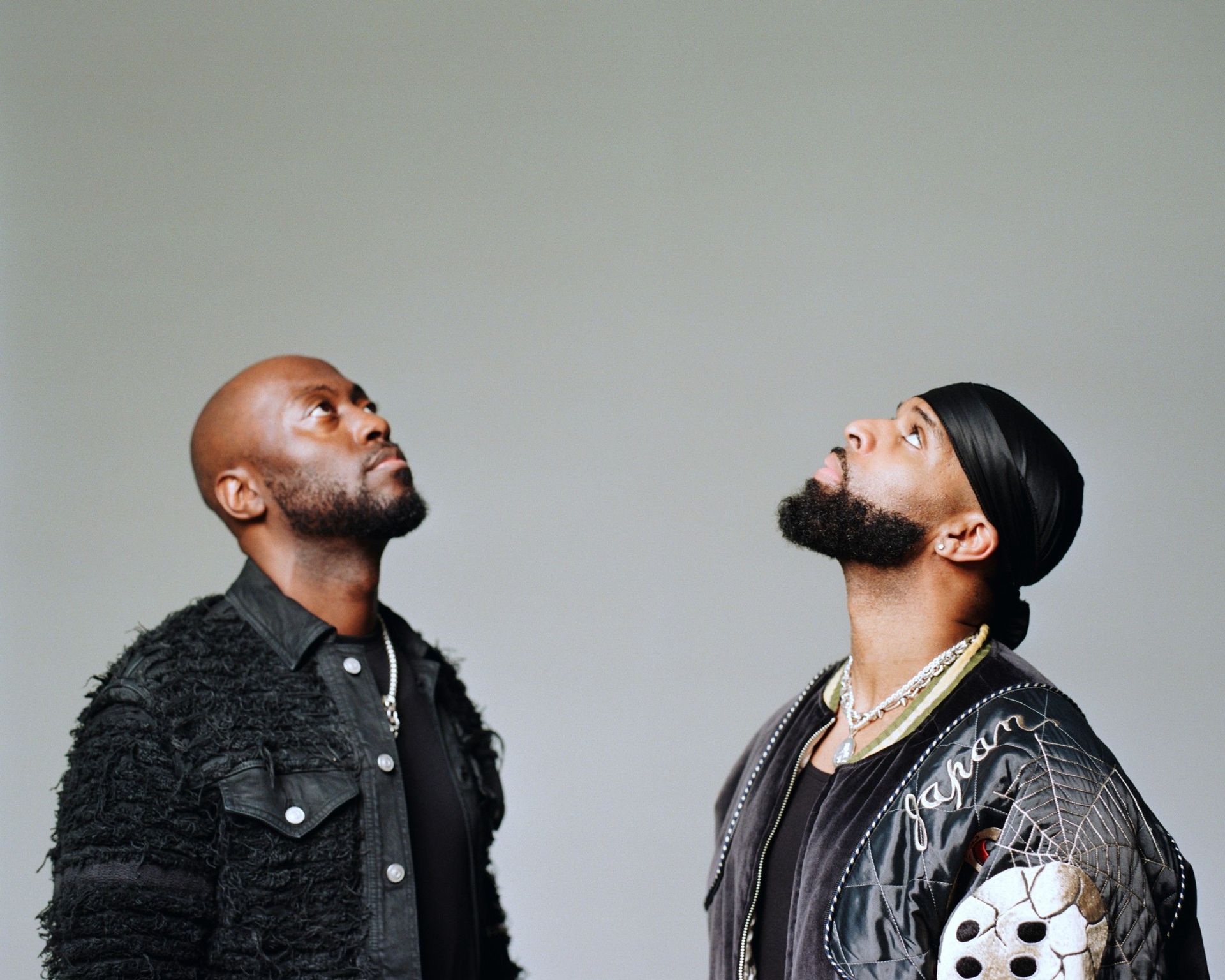 Two men in dark jackets looking upward against a plain gray background.