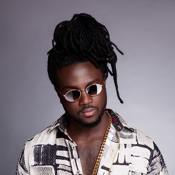 Portrait of a person with sunglasses, dark dreadlocks, and a patterned shirt against a gray background