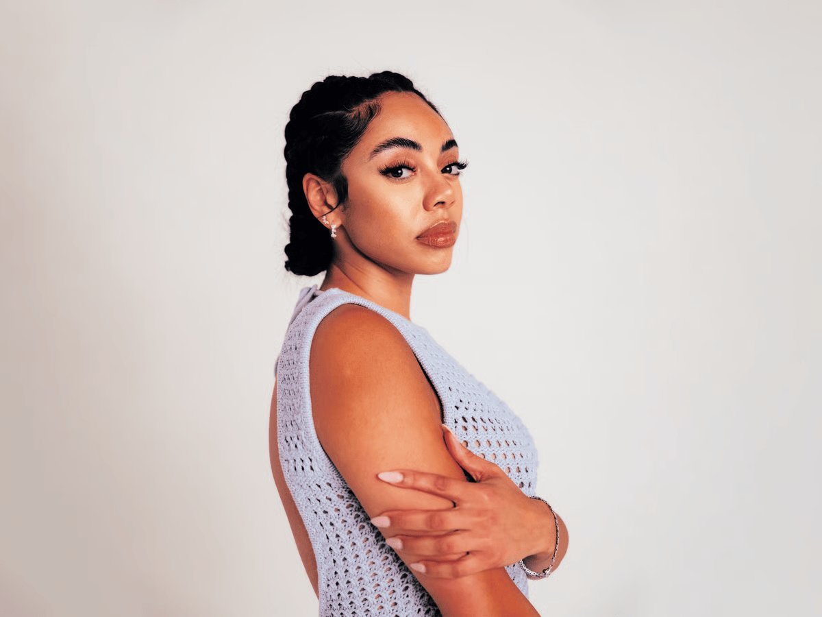 Portrait of a woman in a light lavender sleeveless top, standing in profile with arms crossed against a plain background