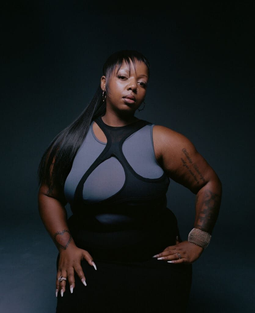 Portrait of a person in a black outfit against a dark studio backdrop, seated with a neutral expression