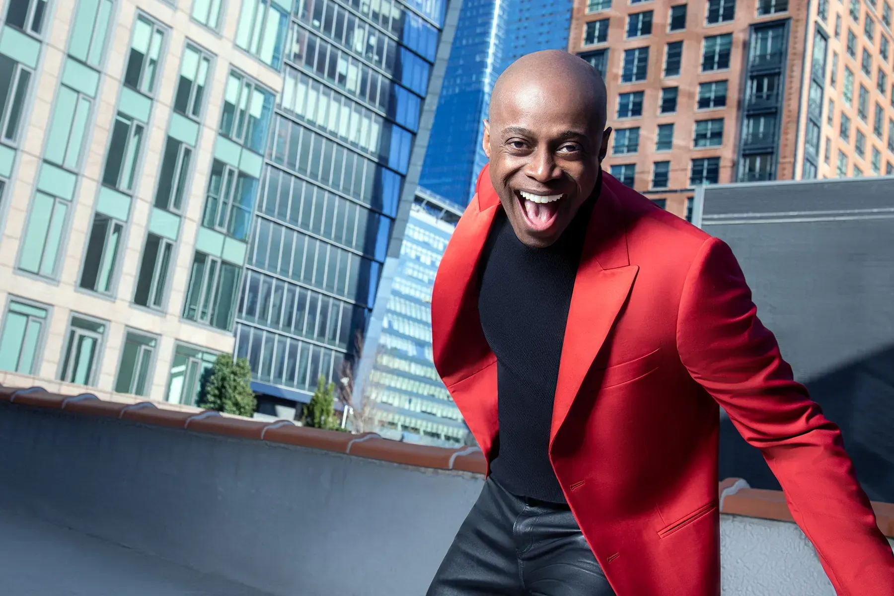 Smiling person in a red blazer leaning forward on a rooftop with city buildings in the background