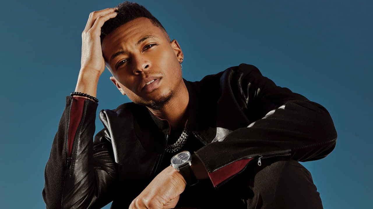 Person in a black jacket sitting against a blue background, resting one hand on their head.