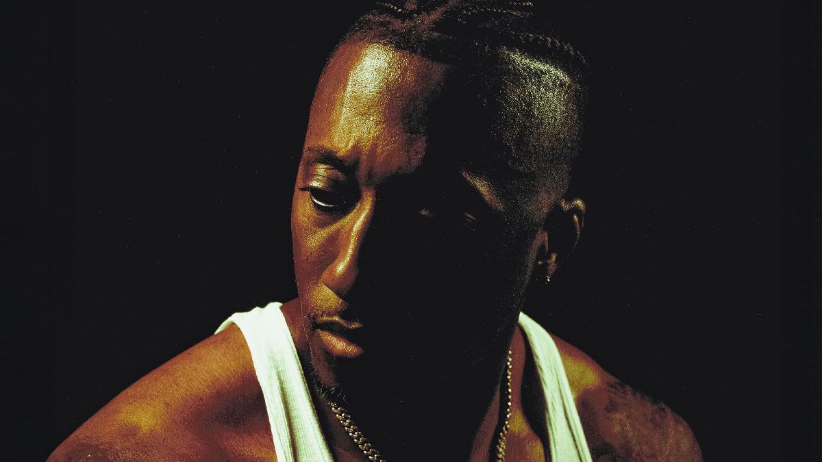 Close-up portrait of a person in a white tank top, looking down against a dark background.